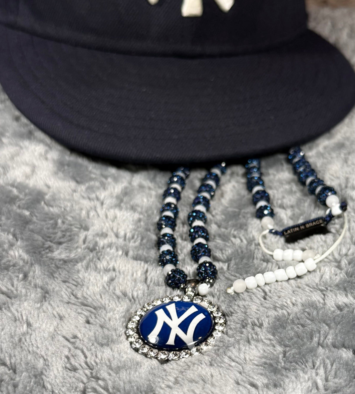A close-up of a New York Yankees beaded necklace with a blue and white logo pendant, displayed next to a matching baseball cap on a gray textured surface.